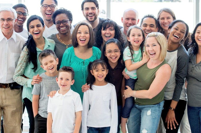 A multi-ethnic family group standing together happily while smiling and looking at the camera.